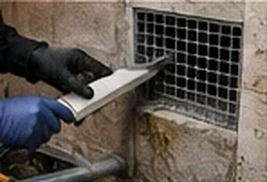 Gloved hands working on a vent opening in a masonry wall (illustrative image)